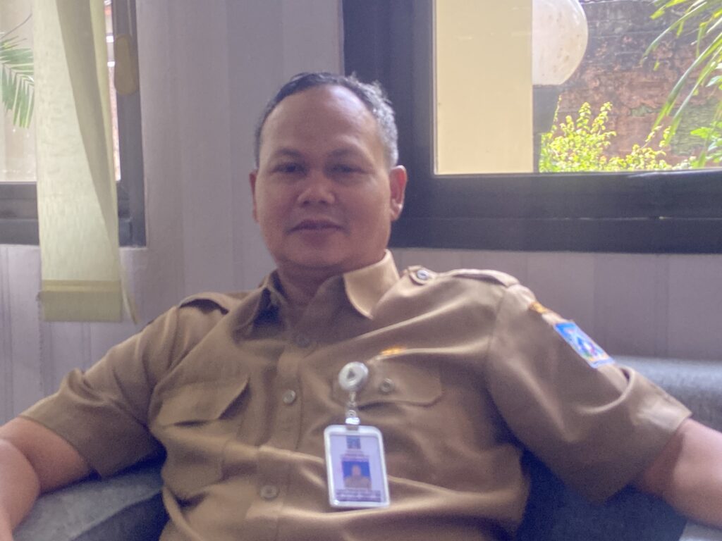 Man in khaki uniform wearing an ID badge, seated indoors near a window.