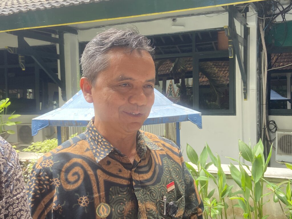 IMG_6309 - Mabur.co Man in a patterned batik shirt outdoors, near green and white building with plants in the foreground.