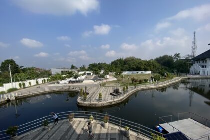 Wide canal with a curved pedestrian promenade, potted plants, and people walking along low railings under a clear blue sky; a circular island feature sits in the water with trees nearby to the left-center.