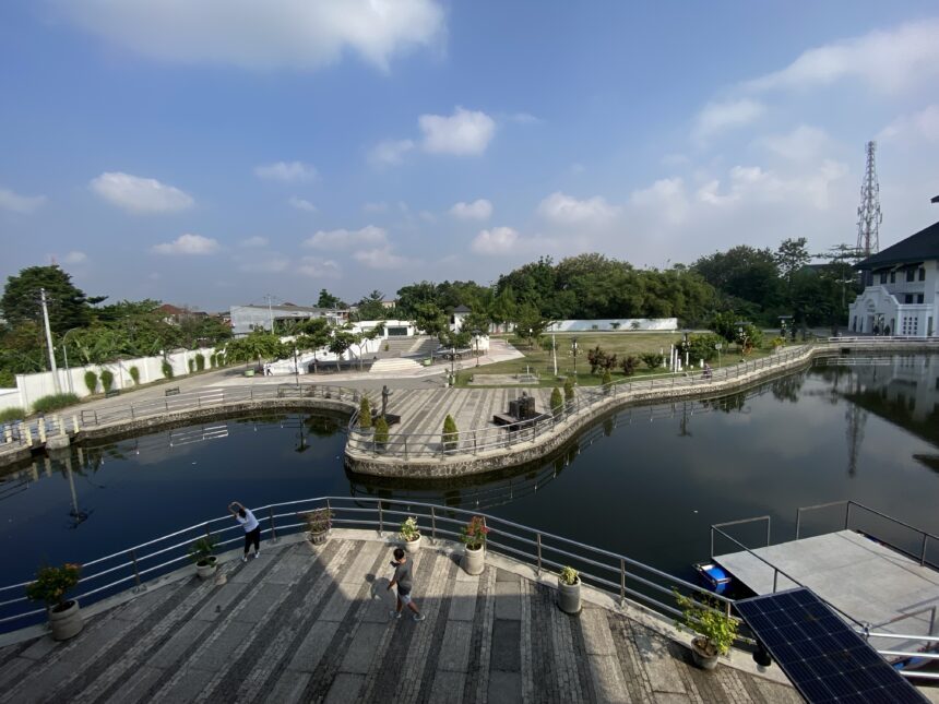 Wide canal with a curved pedestrian promenade, potted plants, and people walking along low railings under a clear blue sky; a circular island feature sits in the water with trees nearby to the left-center.