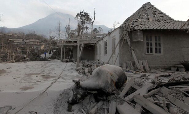 Destroyed village street with collapsed roofs, rubble, and distant mountains; signs of severe disaster.
