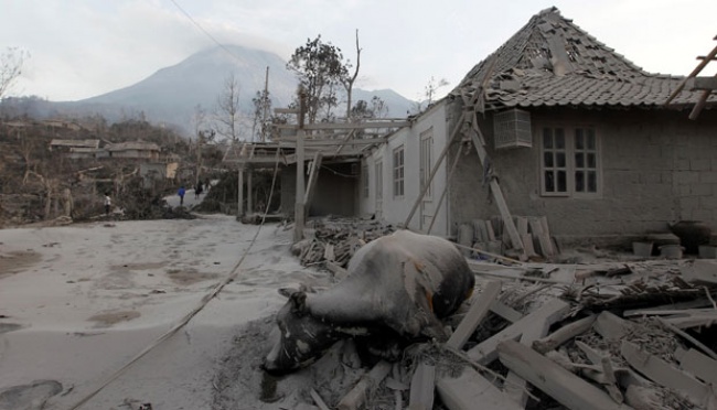 Destroyed village street with collapsed roofs, rubble, and distant mountains; signs of severe disaster.