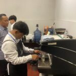 Barista wearing apron operates espresso machine while colleagues watch in a cafe setup with a water cooler and cups nearby.