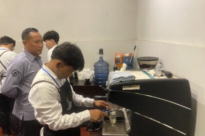 Barista wearing apron operates espresso machine while colleagues watch in a cafe setup with a water cooler and cups nearby.