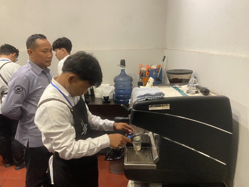 Barista wearing apron operates espresso machine while colleagues watch in a cafe setup with a water cooler and cups nearby.