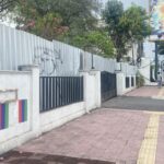 Sidewalk along a white wall with a black metal fence, pink flowering trees, and a distant person walking; colorful banners decorate the lower wall and a billboard is visible nearby.