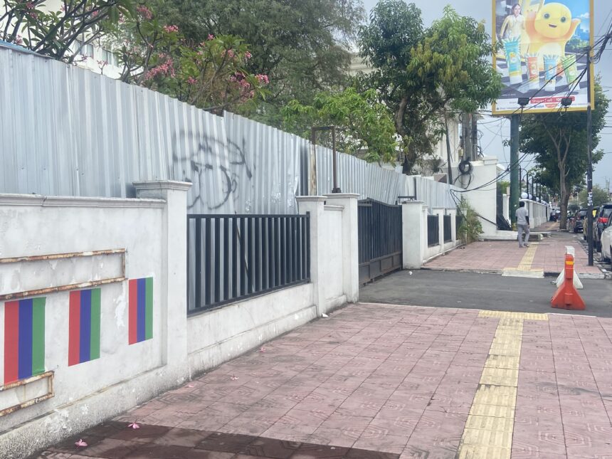 Sidewalk along a white wall with a black metal fence, pink flowering trees, and a distant person walking; colorful banners decorate the lower wall and a billboard is visible nearby.