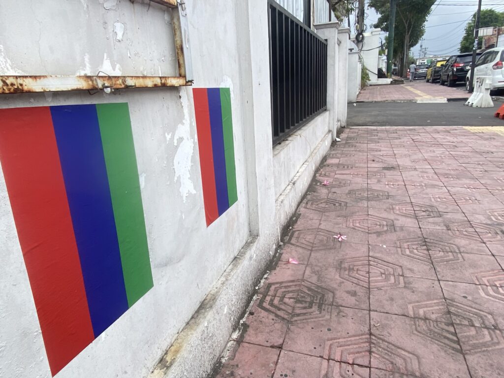White wall decorated with bold vertical red, blue, and green stripes beside a sidewalk and row of parked cars down the street.