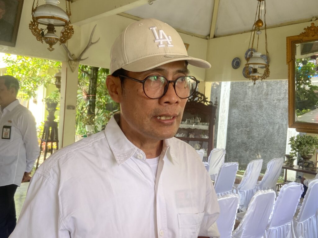 IMG_6490 - Mabur.co Man wearing beige Los Angeles cap and black-rimmed glasses at an indoor event, white chair covers in background.