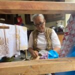 Older woman with glasses sits at a wooden loom, weaving patterned fabric as colorful textiles hang nearby, with a person in a red headscarf in the background.