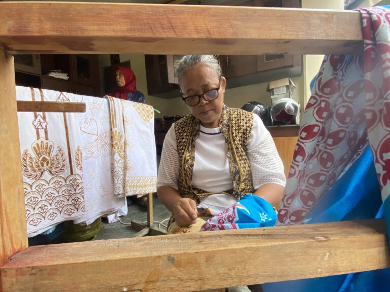 Older woman with glasses sits at a wooden loom, weaving patterned fabric as colorful textiles hang nearby, with a person in a red headscarf in the background.