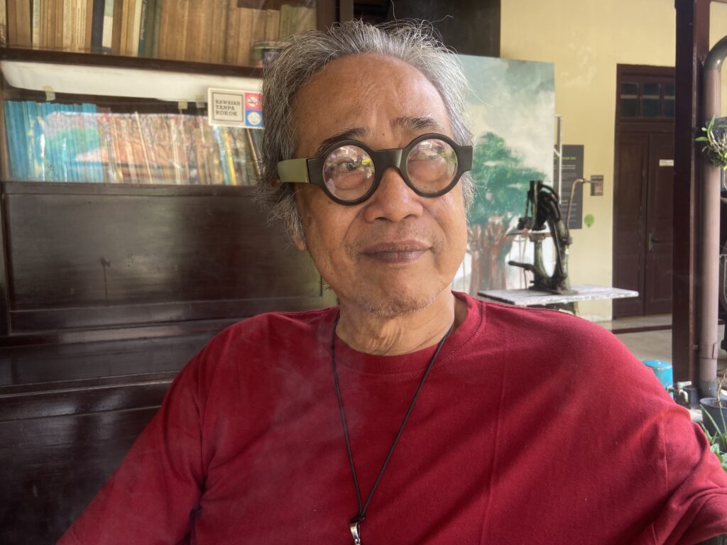 IMG_6591 - Mabur.co Man wearing oversized round glasses indoors, with bookshelves visible behind him and a red shirt.
