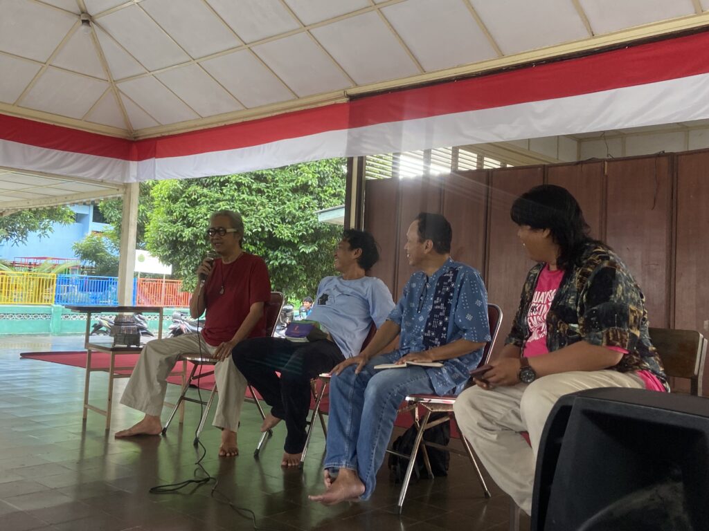 IMG_6595 - Mabur.co Group of people seated on chairs under a red-and-white canopy, one person holding a microphone and speaking