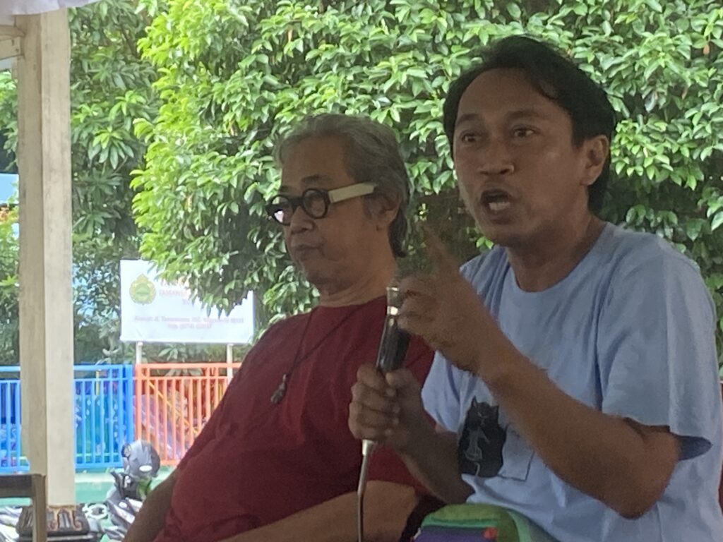 IMG_6615 - Mabur.co Two men sit outside, one in a red shirt with glasses and the other in a light blue shirt, speaking or presenting with greenery and colorful railings in the background.