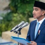 Man in a blue suit and black songkok delivering a speech at a podium with two microphones, holding a blue folder.