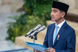 Man in a blue suit and black songkok delivering a speech at a podium with two microphones, holding a blue folder.