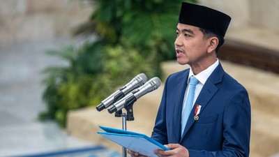 Man in a blue suit and black songkok delivering a speech at a podium with two microphones, holding a blue folder.
