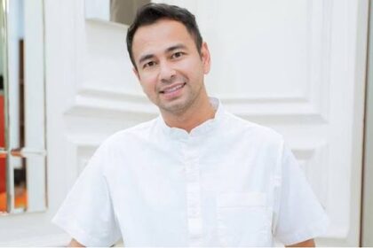 Smiling man in a white short-sleeve shirt posing outdoors beside a light-colored building.