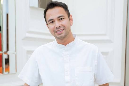 Smiling man in a white short-sleeve shirt posing outdoors beside a light-colored building.