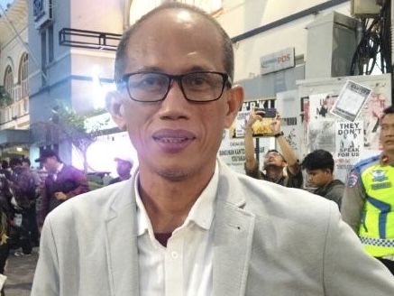 Man wearing glasses and a light gray blazer smiles at the camera in a crowded street event at night, with signs and a security officer in the background.