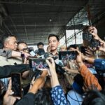 Man in a batik shirt speaks to reporters crowded around him, microphones and smartphones pointed in from all sides in an indoor venue.
