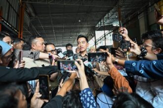 Man in a batik shirt speaks to reporters crowded around him, microphones and smartphones pointed in from all sides in an indoor venue.