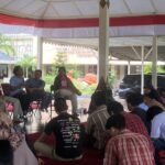 Person holding a microphone and speaking to a small audience seated in a semicircle under a red-and-white canopy outdoors, with plants in the background.