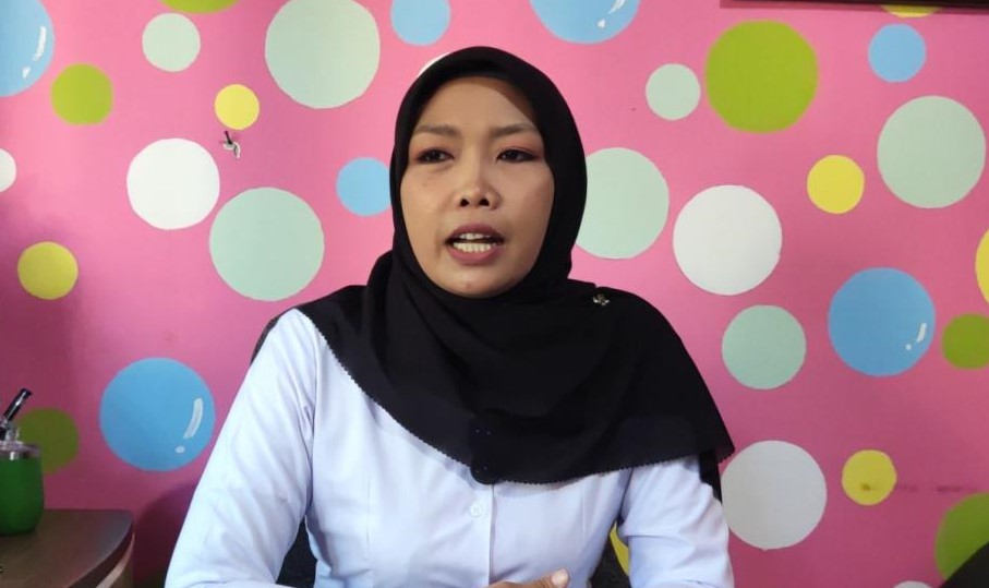 IMG_6789 - Mabur.co Woman in a black hijab and white blouse speaks in front of a bright pink wall with colorful circles.