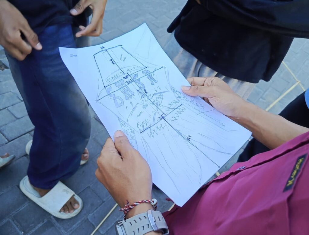 IMG_6799jpg - Mabur.co Two people hold a hand-drawn schematic or diagram on a sheet of white paper outdoors, showing a rough building layout with labeled sections.