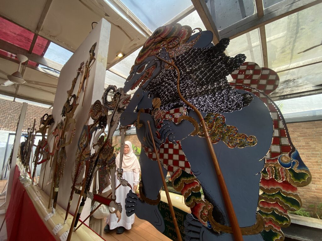 Row of colorful traditional shadow puppets on sticks displayed in a shop, featuring intricate patterns and scales, with a woman in a white hijab visible in the background.