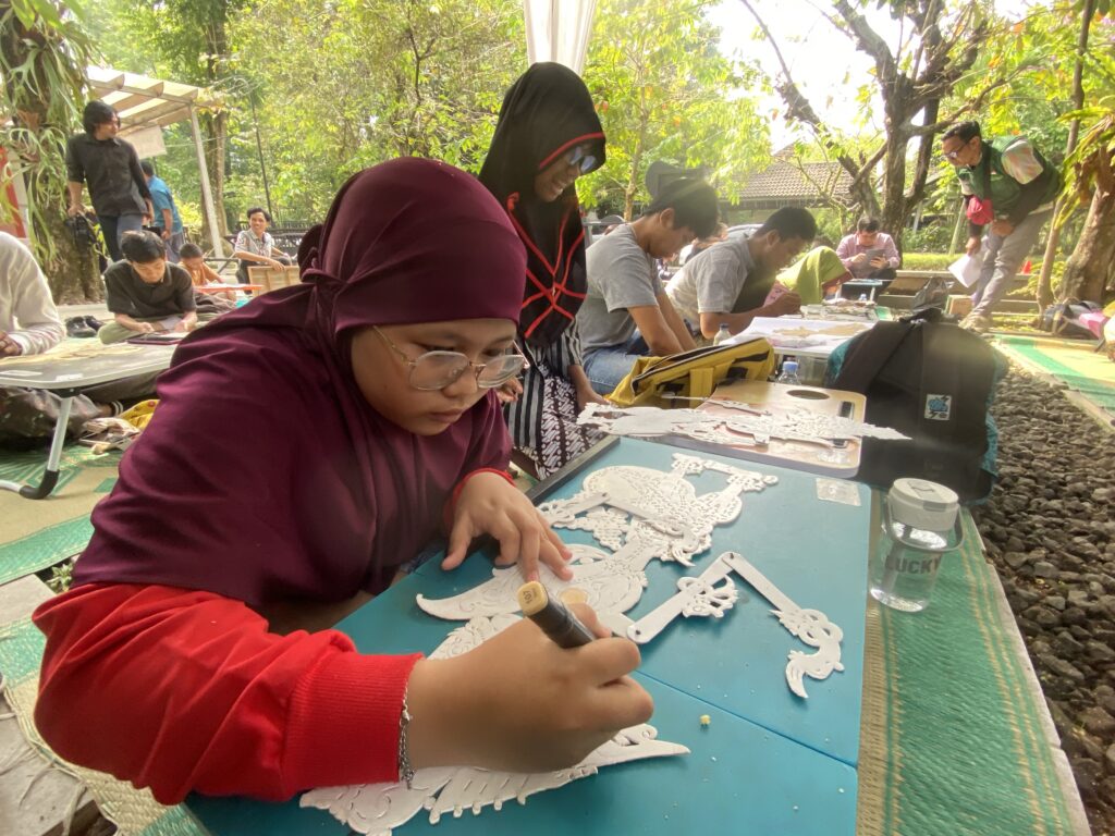 Woman in a maroon hijab and red sleeves works on white paper lace decorations at an outdoor craft table, surrounded by other crafters under trees.