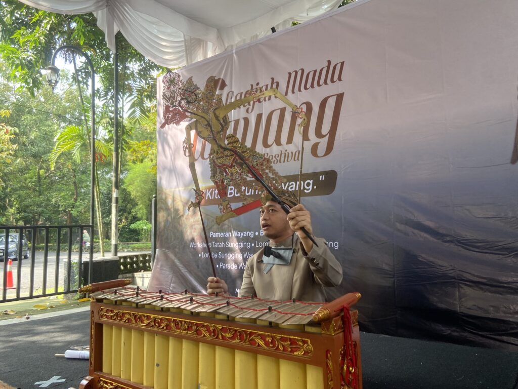 Performer in traditional attire plays a decorated gamelan instrument on an outdoor stage with a festival banner behind.