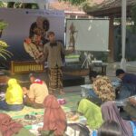 Performer in traditional Indonesian attire holding carved wayang puppets for a children’s puppet workshop outdoors under a canopy.