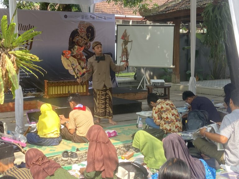 Performer in traditional Indonesian attire holding carved wayang puppets for a children’s puppet workshop outdoors under a canopy.