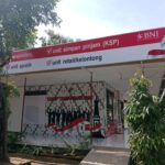 Outdoor bank kiosk sign reading 'unit simpan pinjam (KSP)' with BNI logo, red banner, and waving red-white ribbon amidst trees