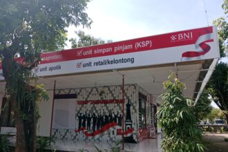 Outdoor bank kiosk sign reading 'unit simpan pinjam (KSP)' with BNI logo, red banner, and waving red-white ribbon amidst trees