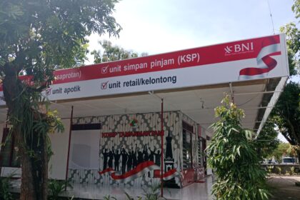 Outdoor bank kiosk sign reading 'unit simpan pinjam (KSP)' with BNI logo, red banner, and waving red-white ribbon amidst trees