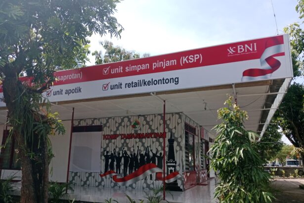 Outdoor bank kiosk sign reading 'unit simpan pinjam (KSP)' with BNI logo, red banner, and waving red-white ribbon amidst trees