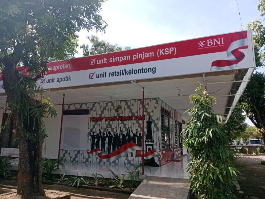 Outdoor bank kiosk sign reading 'unit simpan pinjam (KSP)' with BNI logo, red banner, and waving red-white ribbon amidst trees