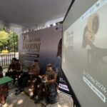 Panel of men in batik shirts seated under a white tent at a Wayang Festival, with a large 'Wayang Festival' banner in the background.