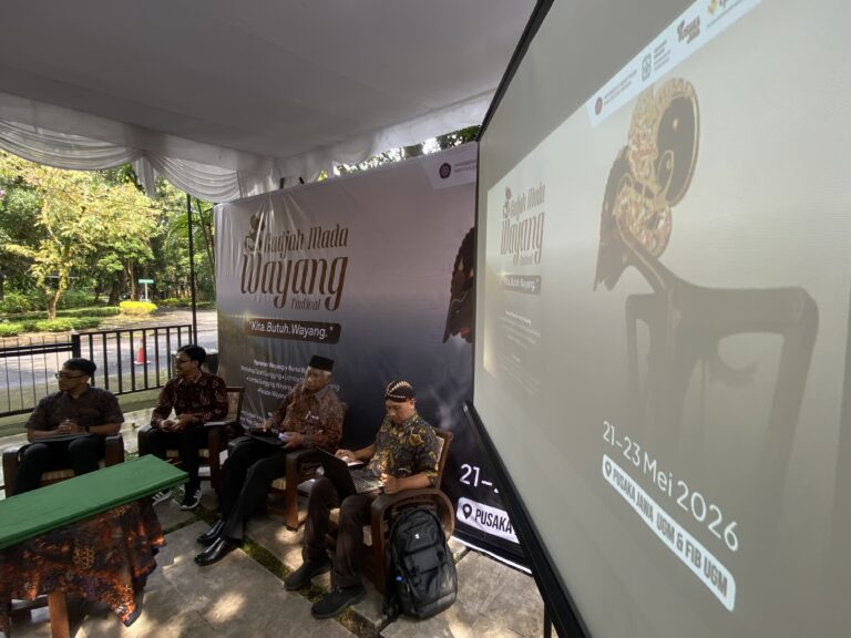 Panel of men in batik shirts seated under a white tent at a Wayang Festival, with a large 'Wayang Festival' banner in the background.