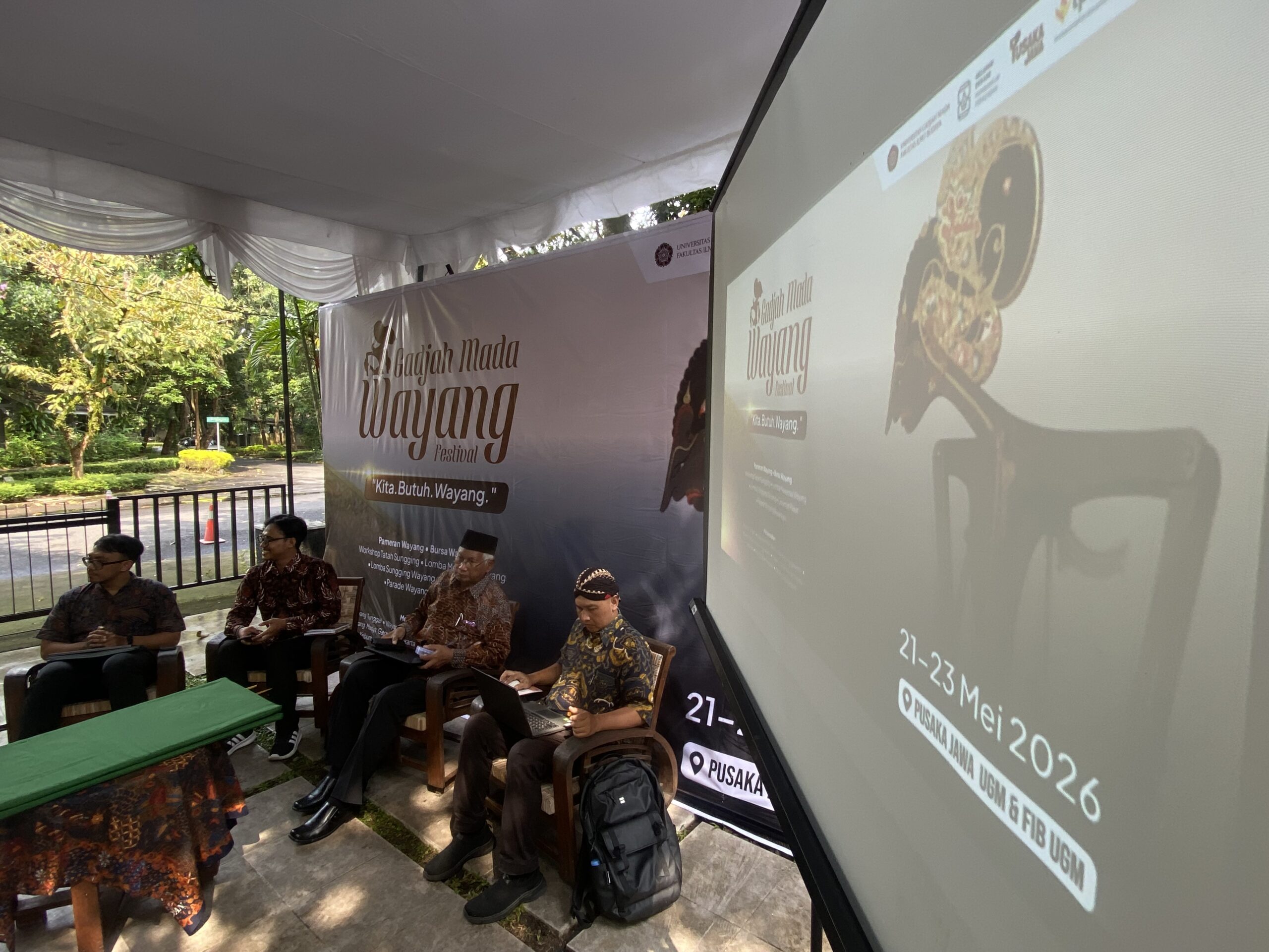 Panel of men in batik shirts seated under a white tent at a Wayang Festival, with a large 'Wayang Festival' banner in the background.