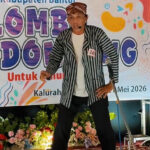 Male performer on a stage with a striped jacket and headset mic, in front of a colorful banner reading 'Lomba Mendongeng' and 'Mei 2026'.