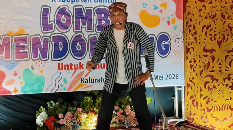 IMG_7025 - Mabur.co Male performer on a stage with a striped jacket and headset mic, in front of a colorful banner reading 'Lomba Mendongeng' and 'Mei 2026'.