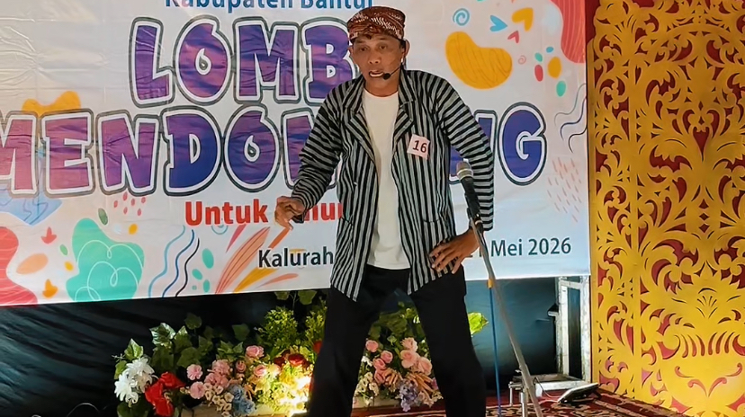 Male performer on a stage with a striped jacket and headset mic, in front of a colorful banner reading 'Lomba Mendongeng' and 'Mei 2026'.