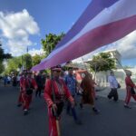 Marchers in red uniforms walk down a street during a parade, holding poles with a large pink and purple striped banner overhead.