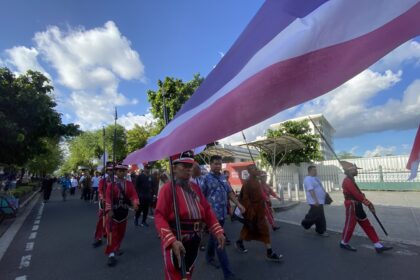 Marchers in red uniforms walk down a street during a parade, holding poles with a large pink and purple striped banner overhead.