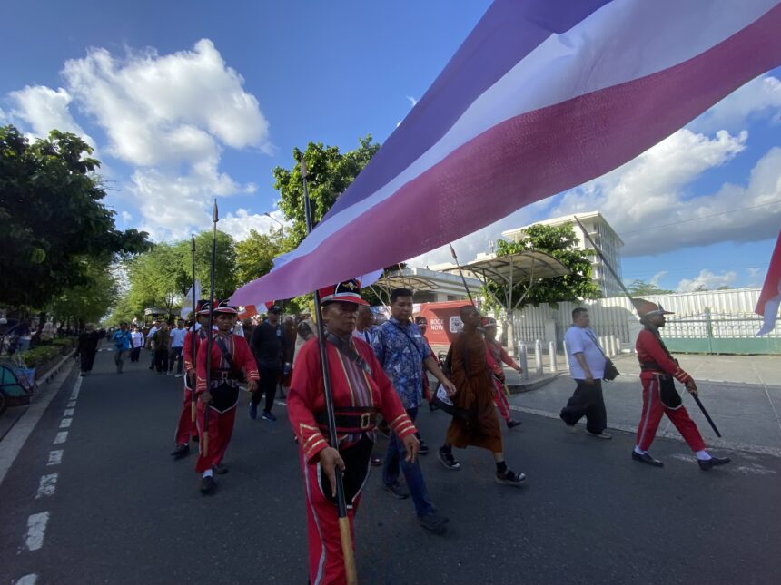 IMG_7170 - Mabur.co Marchers in red uniforms walk down a street during a parade, holding poles with a large pink and purple striped banner overhead.