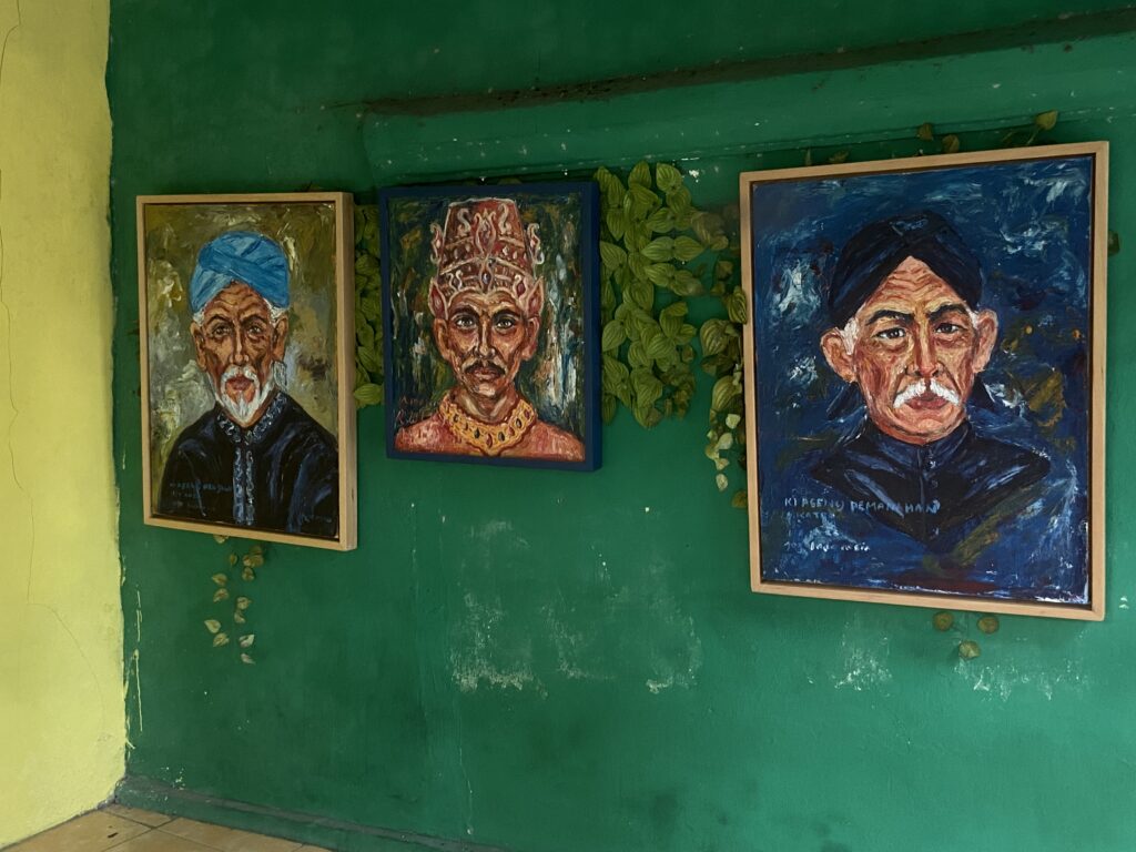 IMG_7290 - Mabur.co Three framed painted portraits of men in traditional clothing hanging on a green wall, with vines climbing between them.