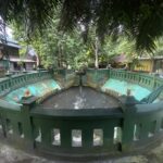 Circular green fountain with a decorative railing and gold finials set in a tropical courtyard, trees overhead and an Indonesian flag nearby